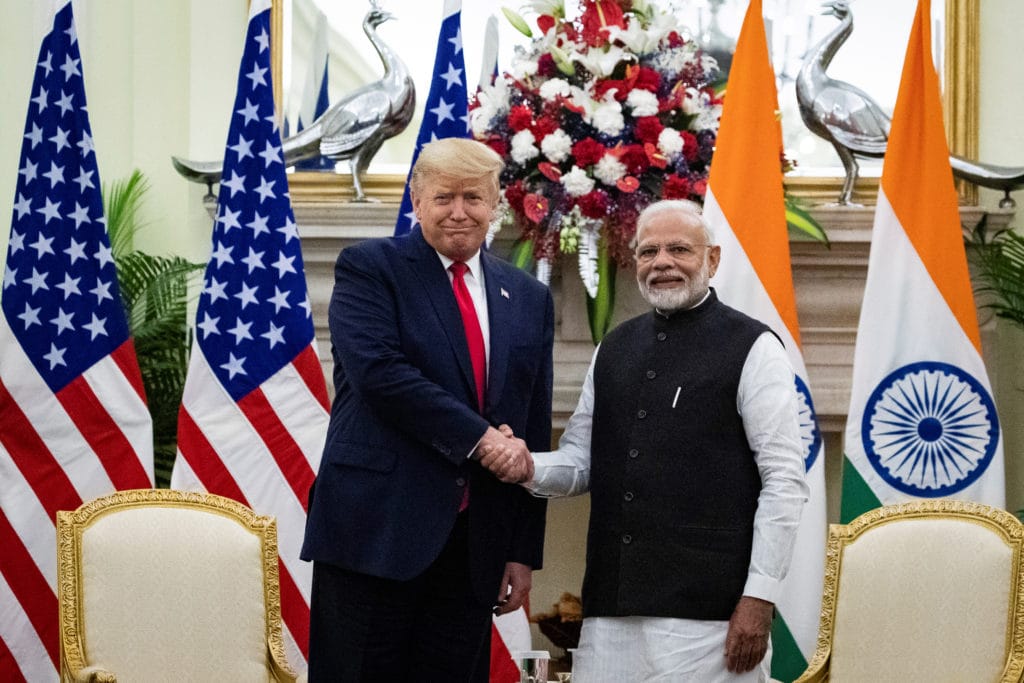 Donald Trump and Narendra Modi shake hands in front of Indian and U.S. flags during a formal meeting in India.
