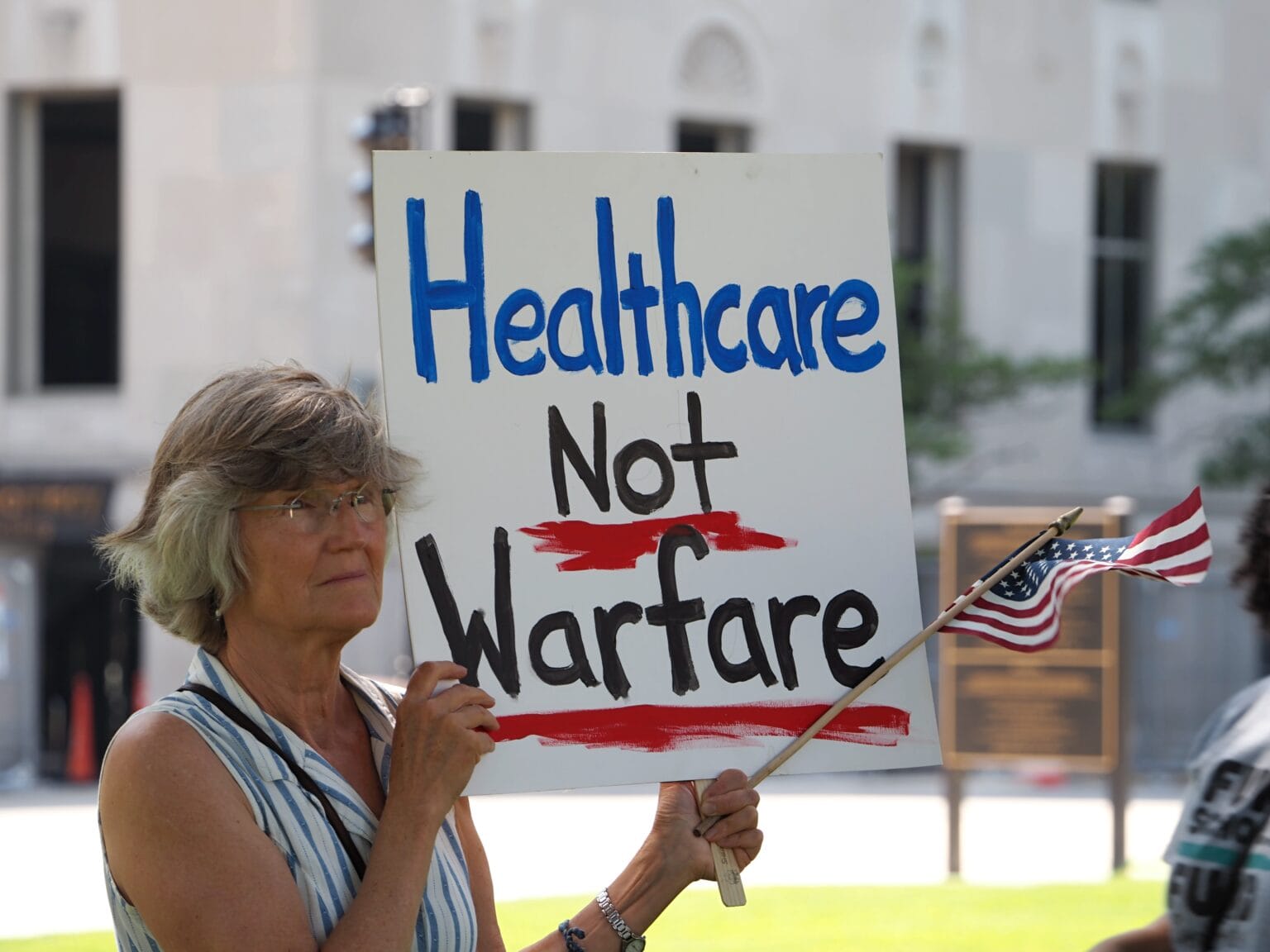 A protester holding a handwritten cardboard sign reading “Healthcare Not Warfare”