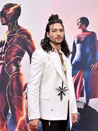 Ezra Miller dressed in a white suit at a premiere event with The Flash poster in the background featuring DC superheroes.