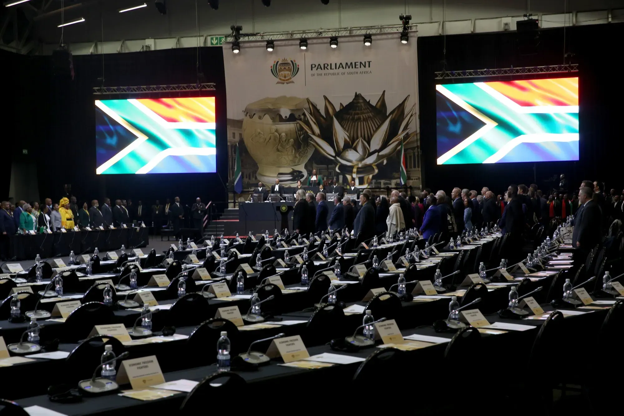 Rows of labeled seats and water bottles in South Africa’s Parliament hall ahead of session, flanked by digital South African flags.