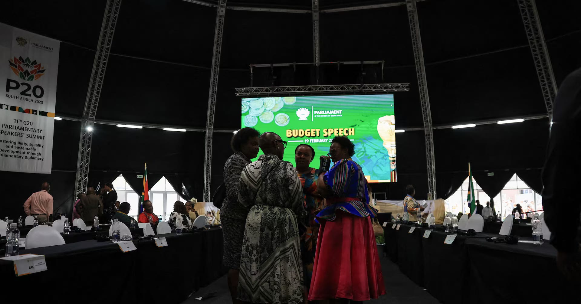 Wide view of South Africa’s Parliament hall with national flags displayed and members standing for proceedings.