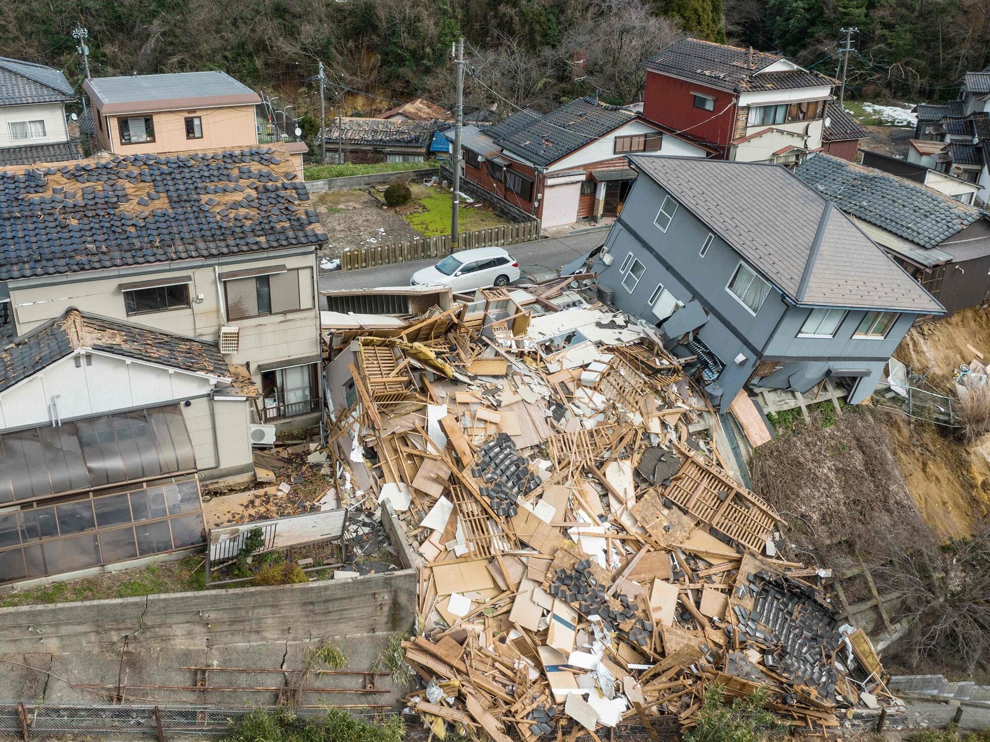 A collapsed wooden house lies in ruins at the edge of a hill, surrounded by intact neighboring buildings in a Japanese town.