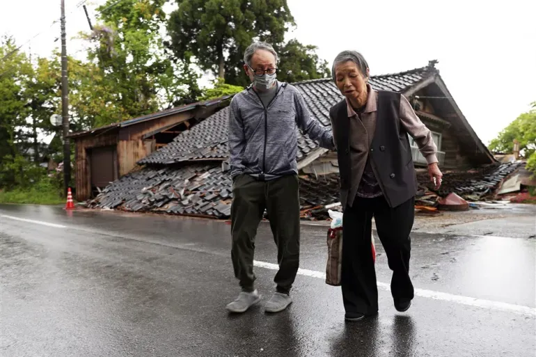 An elderly man helps an older woman walk along a wet road beside a destroyed house with a caved-in tiled roof.