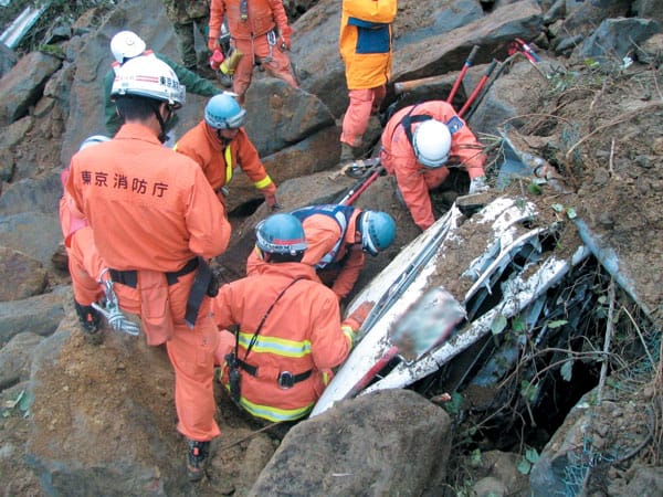 Uniformed Japanese rescue workers inspect the ruins of a destroyed building, surrounded by broken tiles and collapsed beams.