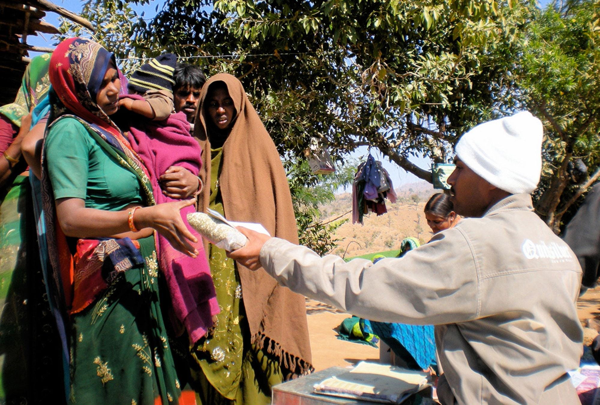 People gather around tables and benches under a canopy at a village vaccination camp.