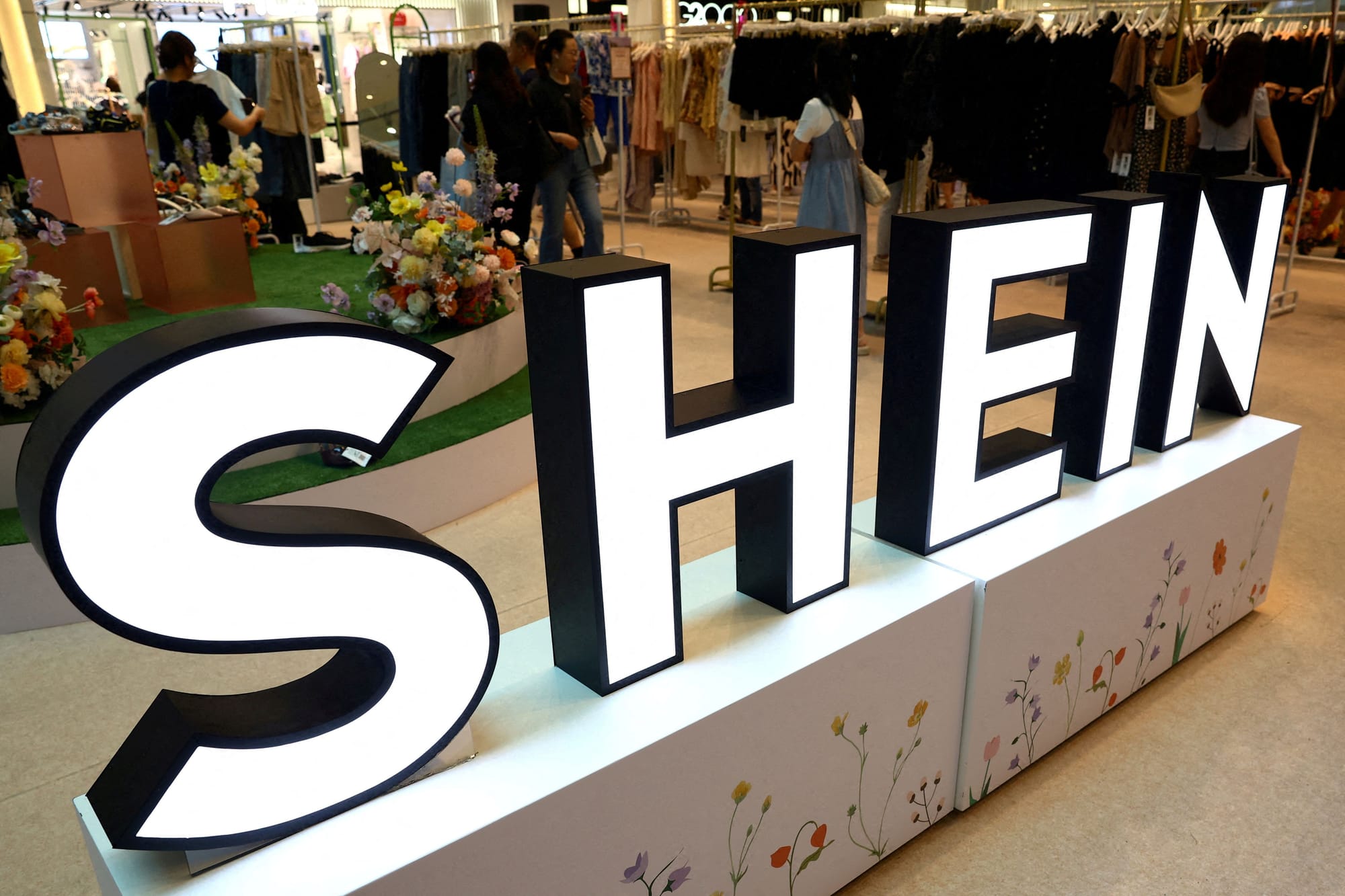 Large illuminated Shein logo at a store surrounded by customers and floral decor.