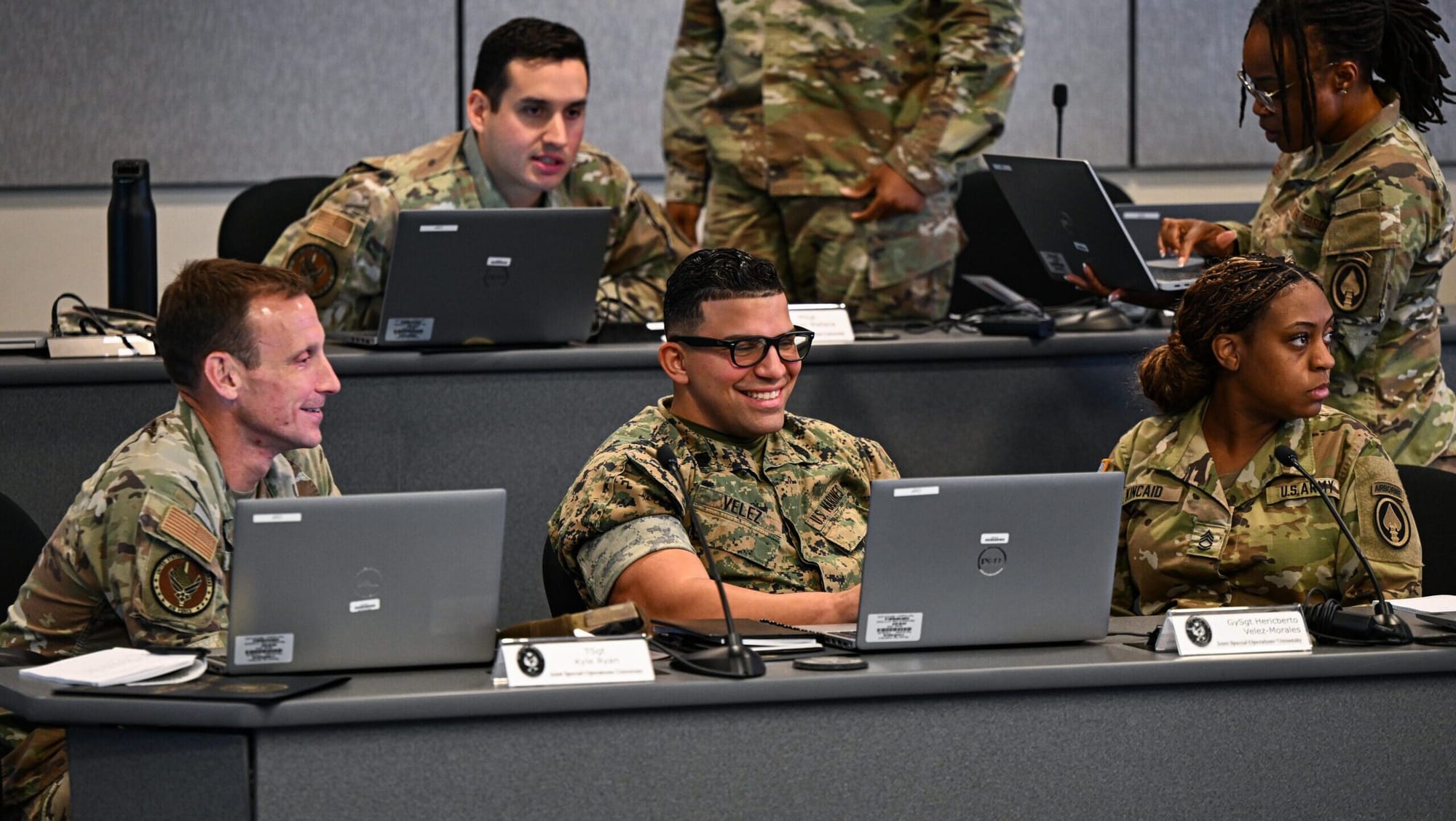 Soldiers gathered around laptops in a military briefing setting.