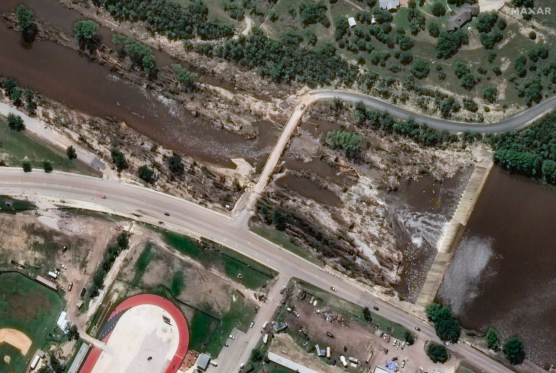 Satellite image showing washed-out debris, damaged vegetation, and a strained spillway near a bridge and highway in Junction, TX.