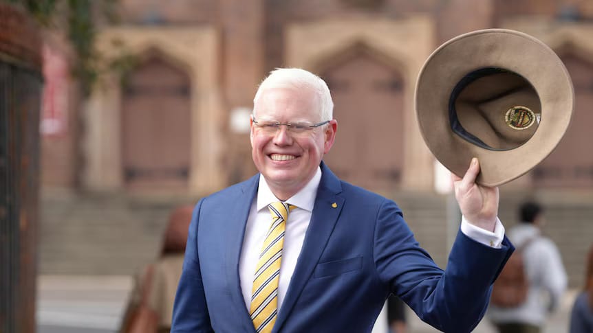 Gareth Ward holding a brown Akubra hat and smiling in a suit and striped tie.