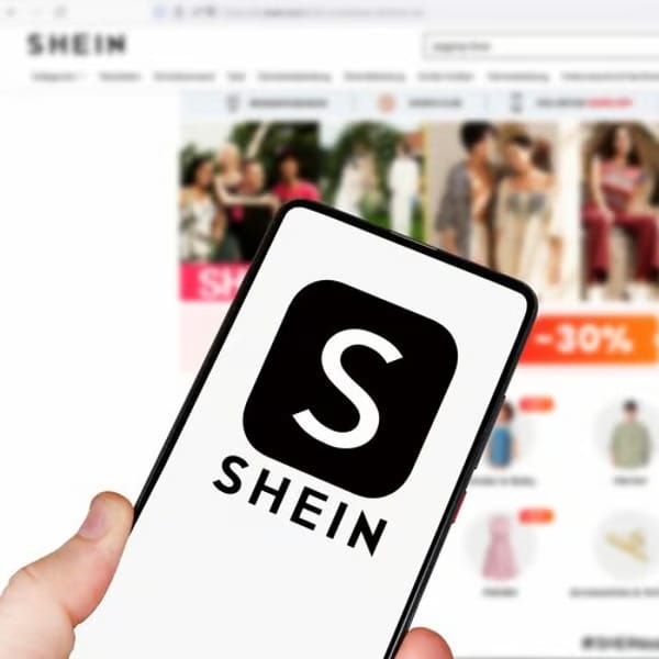 A hand holding a smartphone with the Shein logo on screen, with the Shein online store blurred in the background.