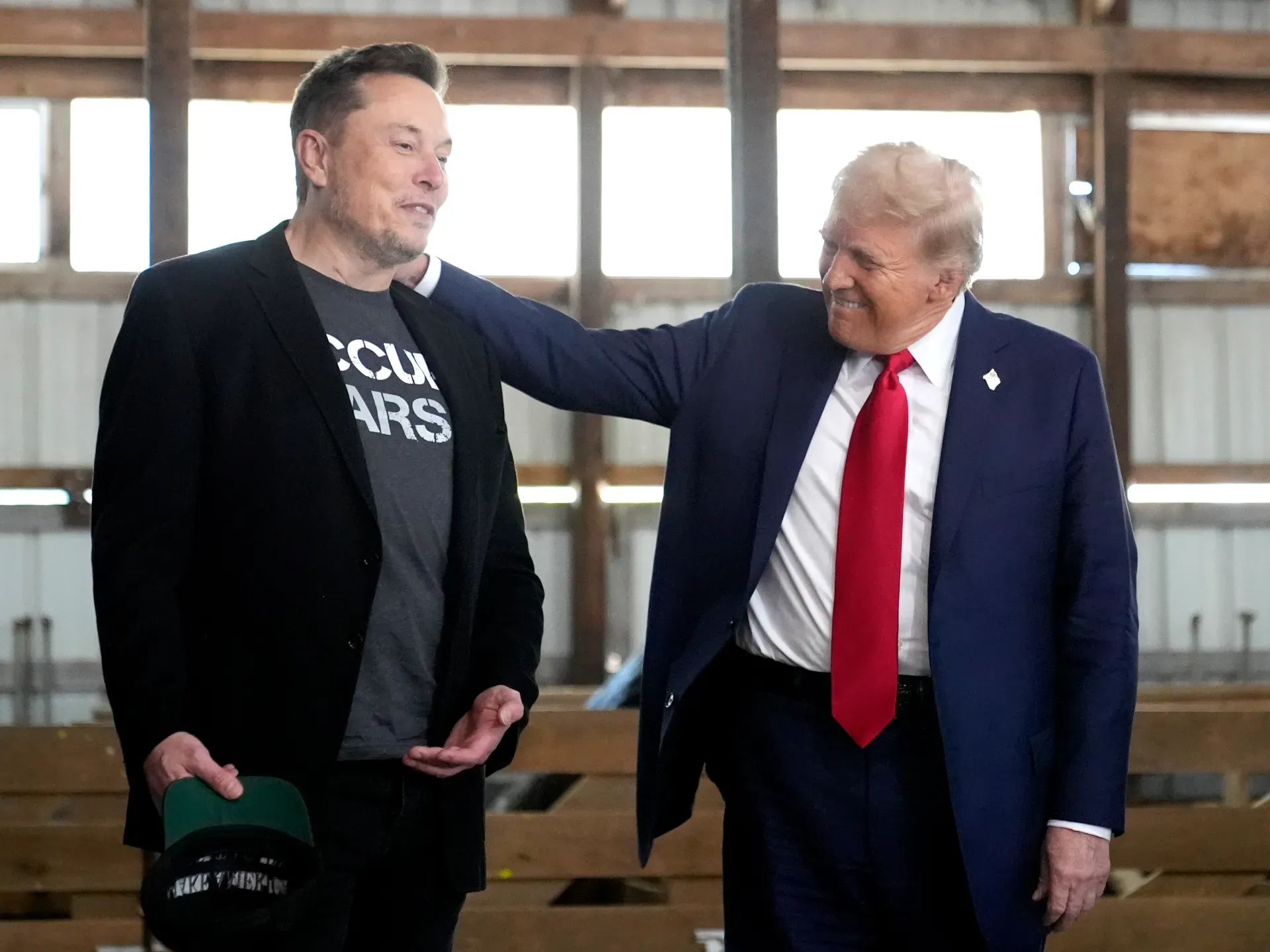 Donald Trump smiles and pats Elon Musk on the shoulder as they stand together inside a wooden barn-like venue, signaling camaraderie.