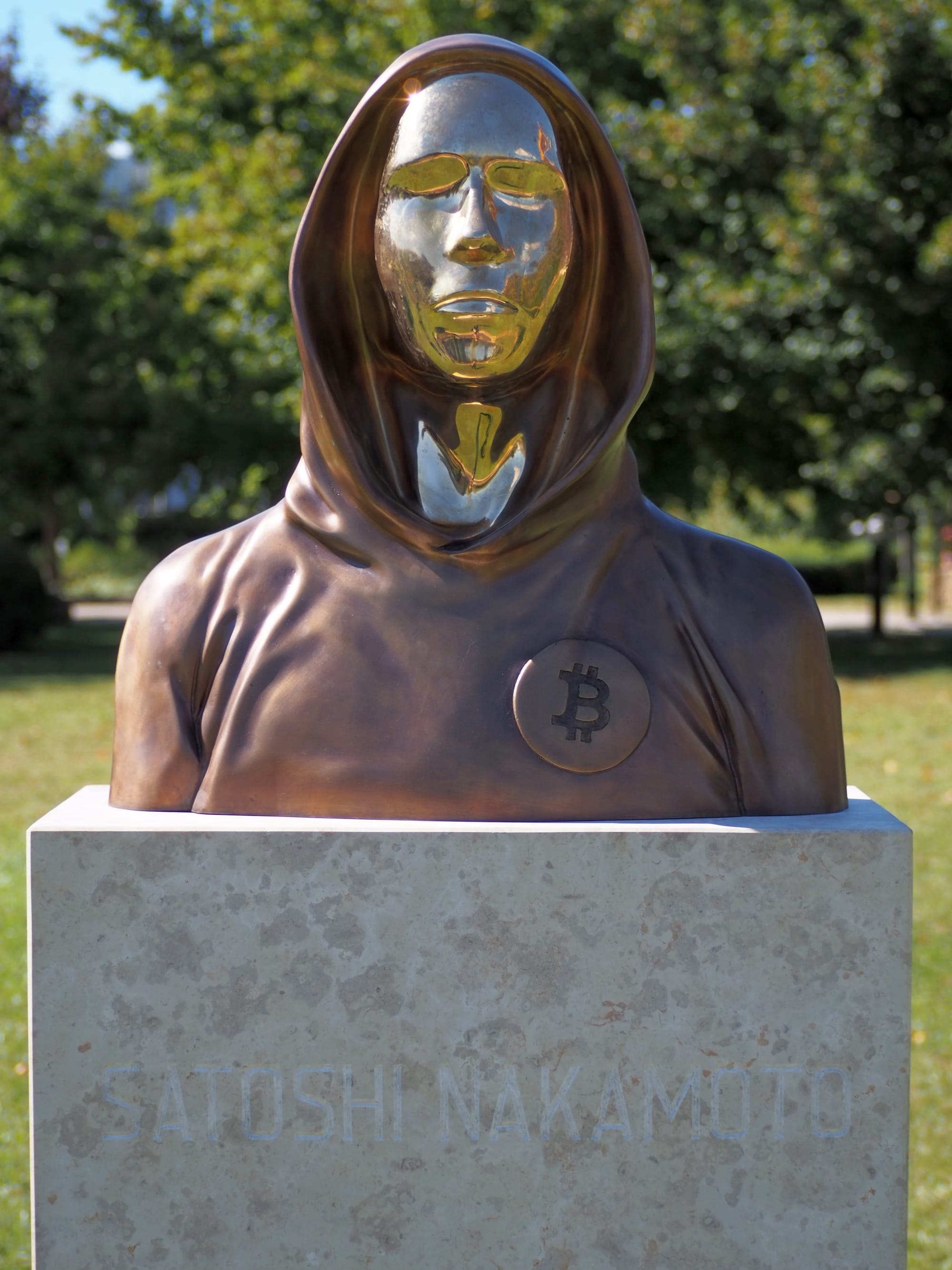 Bronze bust of Satoshi Nakamoto with a hoodie and Bitcoin logo, placed on a stone pedestal with name inscription.