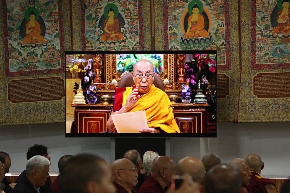 A television screen shows the Dalai Lama delivering a speech, watched by monks seated under traditional Tibetan thangka paintings.
