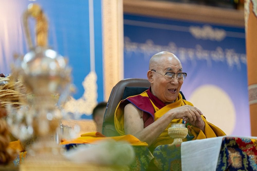 His Holiness the Dalai Lama seated in front of a blue backdrop, smiling during his 90th birthday celebrations.