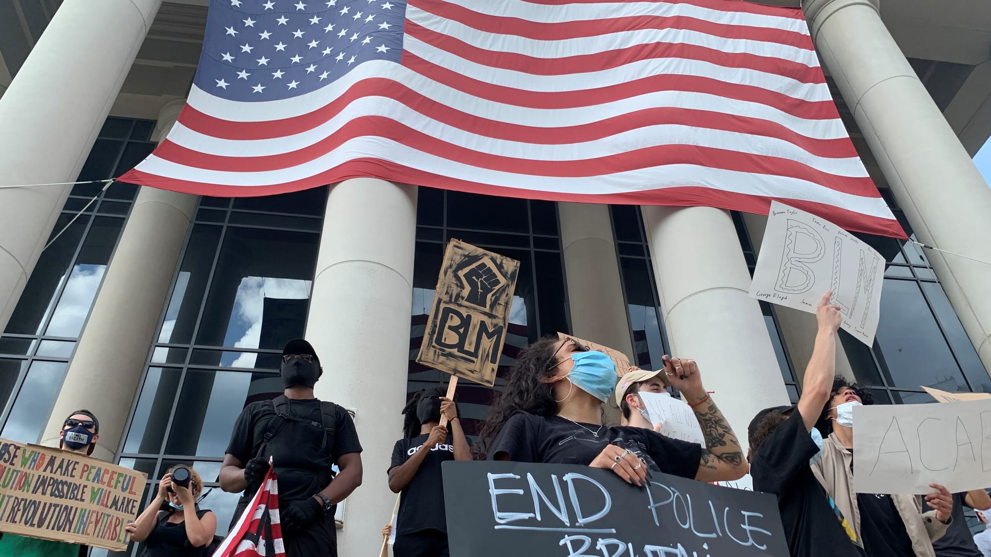 A group of Black Lives Matter protesters holding signs like “END POLICE BRUTALITY” and “BLM” beneath a massive American flag draped over courthouse columns.