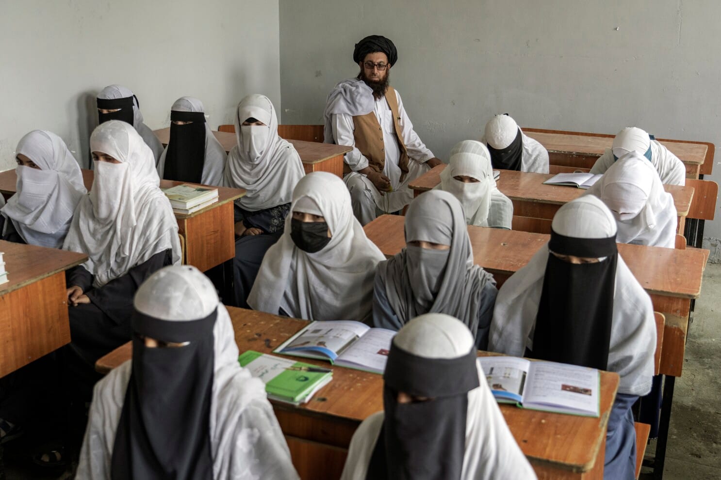 Afghan schoolgirls wearing full-body coverings and niqabs sit in a classroom with a Taliban man monitoring from the back.