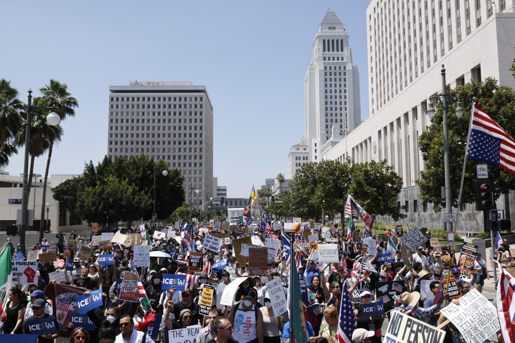 Protesters holding signs and flags march through downtown Los Angeles near federal buildings.