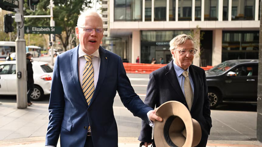 Gareth Ward holding his hat, walking beside a suited man on a city street near Elizabeth St.