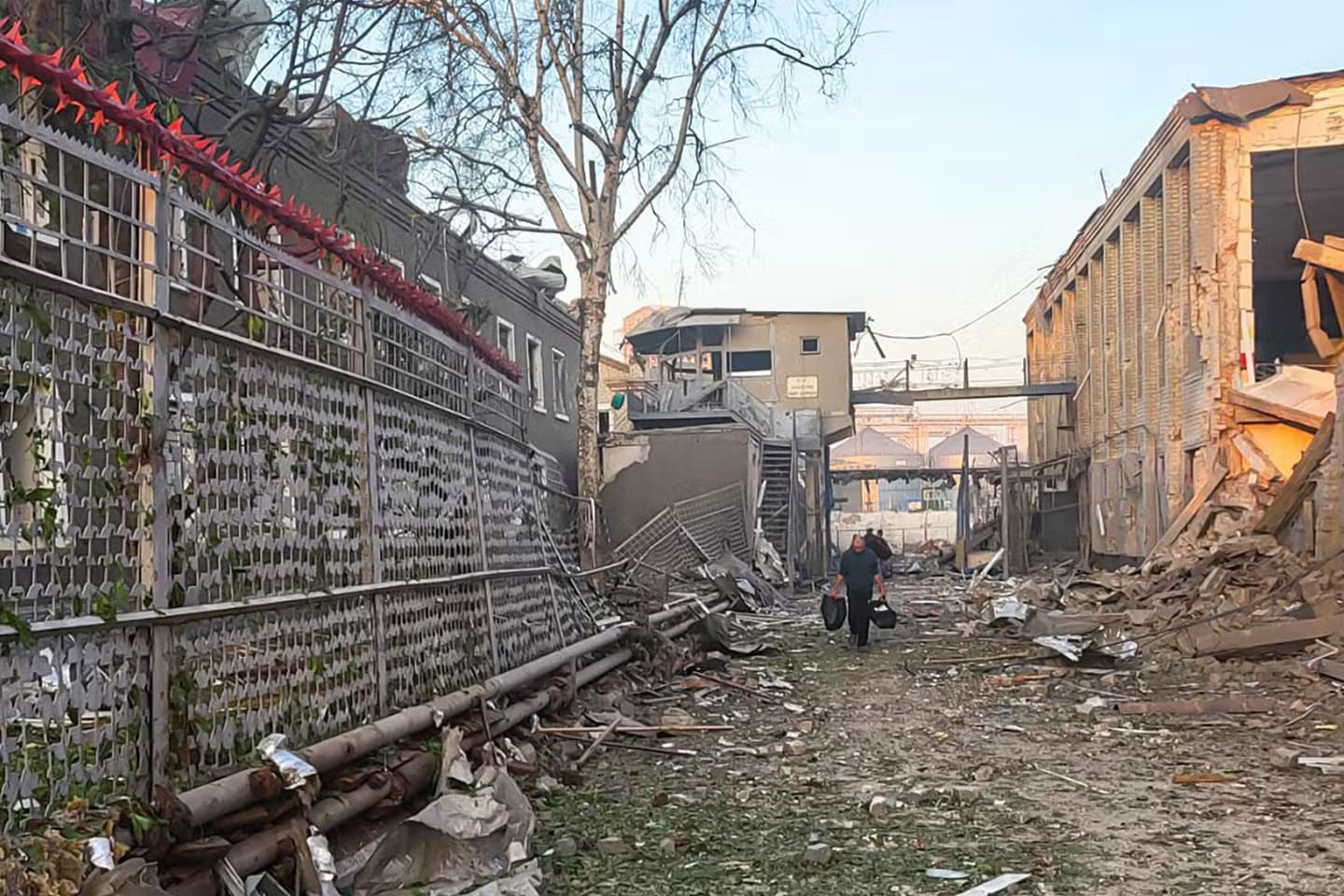 A person carrying bags walks through a destroyed alley surrounded by rubble, shattered buildings, and twisted metal in Kharkiv, Ukraine, following a Russian missile attack.