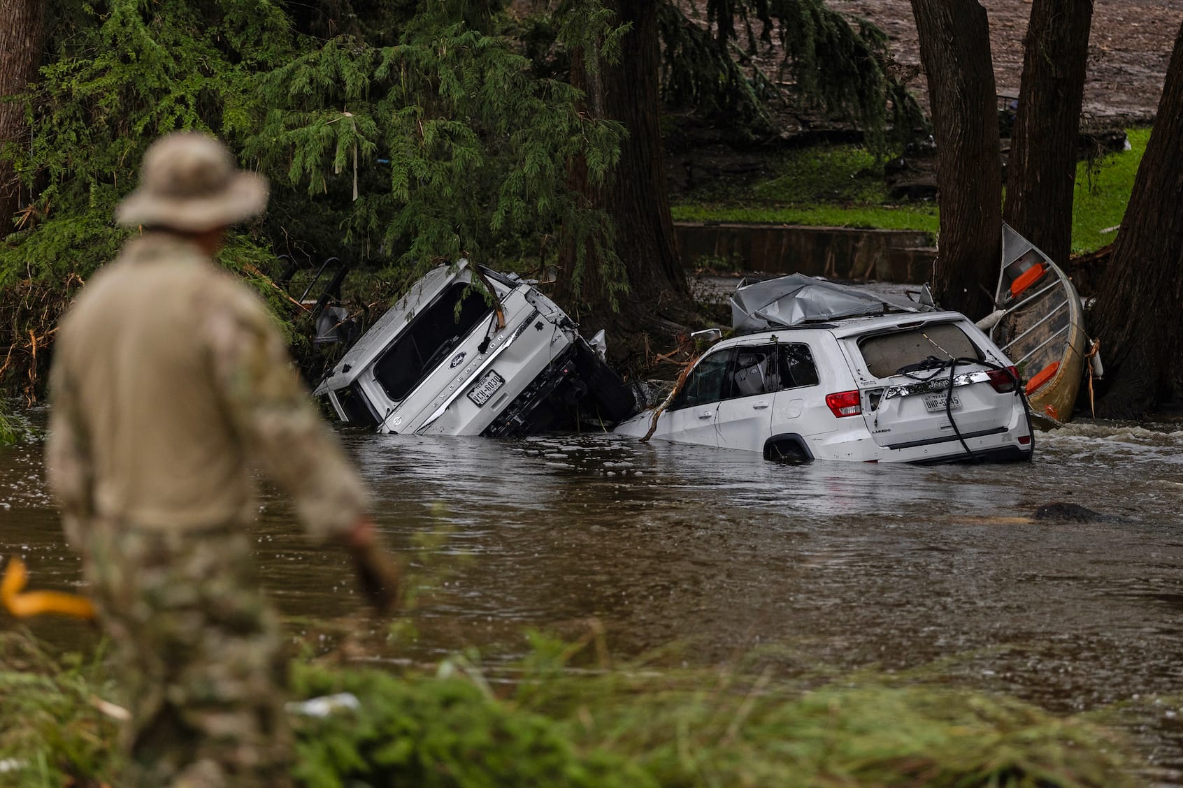 Wrecked cars submerged in floodwater with a rescue worker in the foreground during Texas floods.
