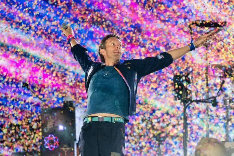 Chris Martin of Coldplay spreads his arms wide while performing on stage, with colorful confetti and lights in the background.