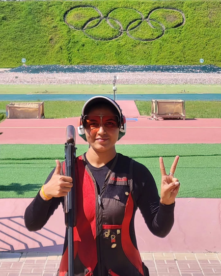Female shooter at an Olympic shooting range holding a shotgun, wearing protective glasses and earmuffs, showing victory signs with both hands in front of the Olympic rings symbol on the grass wall.