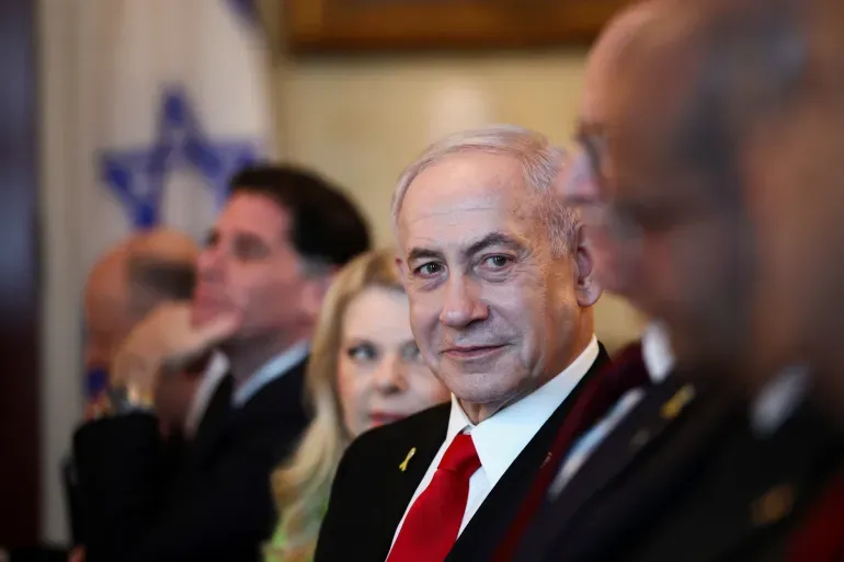 Benjamin Netanyahu sitting among officials, wearing a red tie, with an Israeli flag blurred in the background.