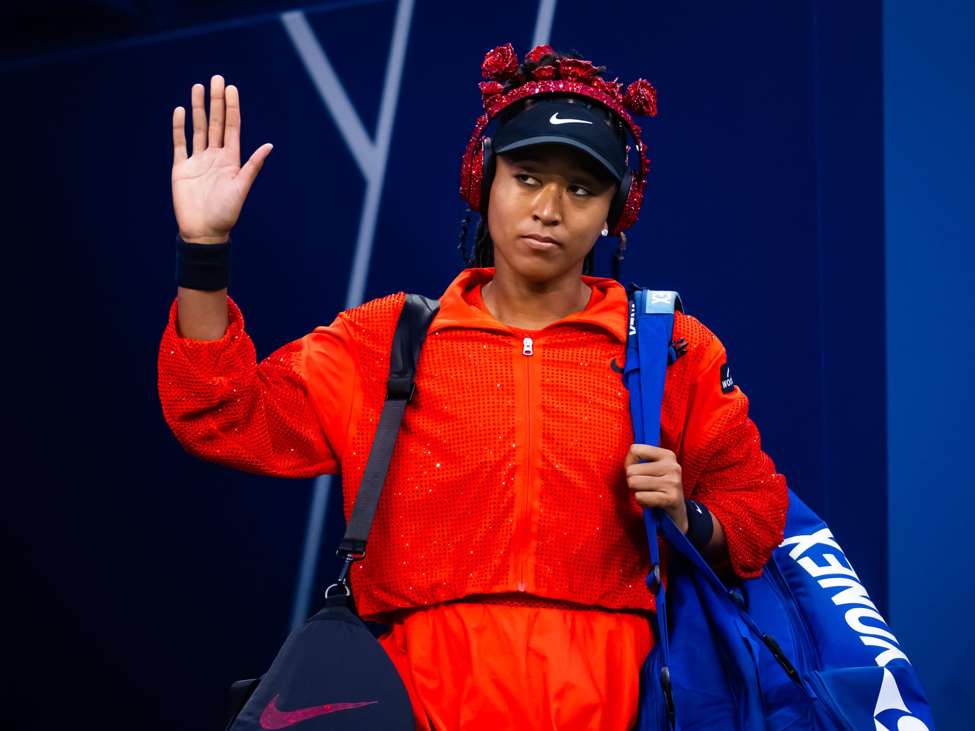 Female tennis player in a sparkling red outfit with braided hair and red headphones waves to the crowd while carrying sports bags at the US Open.