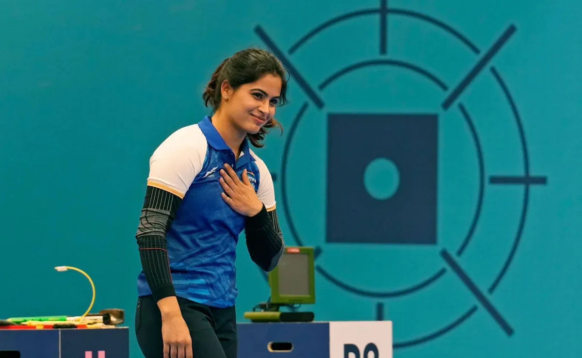 Indian shooter Manu Bhaker smiling with her hand on her chest after a competition, standing in front of a blue shooting target backdrop.