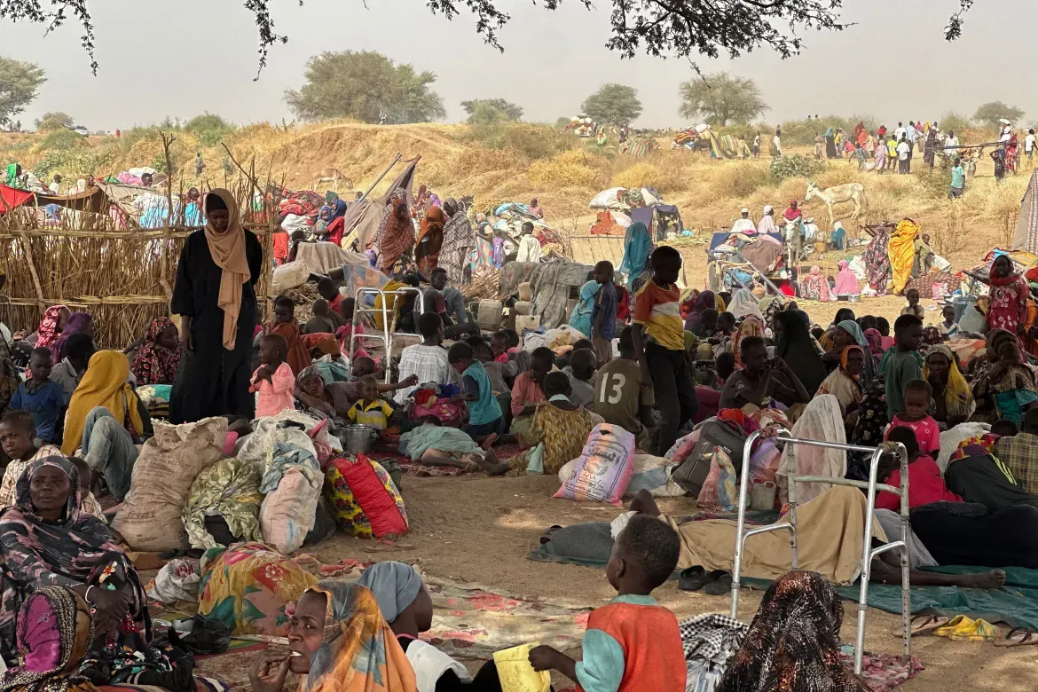 Large crowd of displaced Sudanese families, including women and children, gather under makeshift shelters and trees in a dry rural area near the Chad-Sudan border.