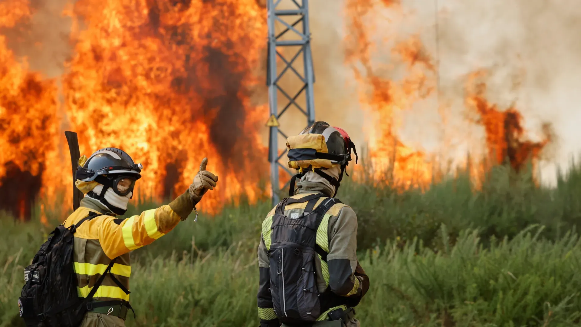 Two firefighters in protective gear stand near raging wildfire flames, with one giving a thumbs-up gesture while intense fire engulfs vegetation in the background.