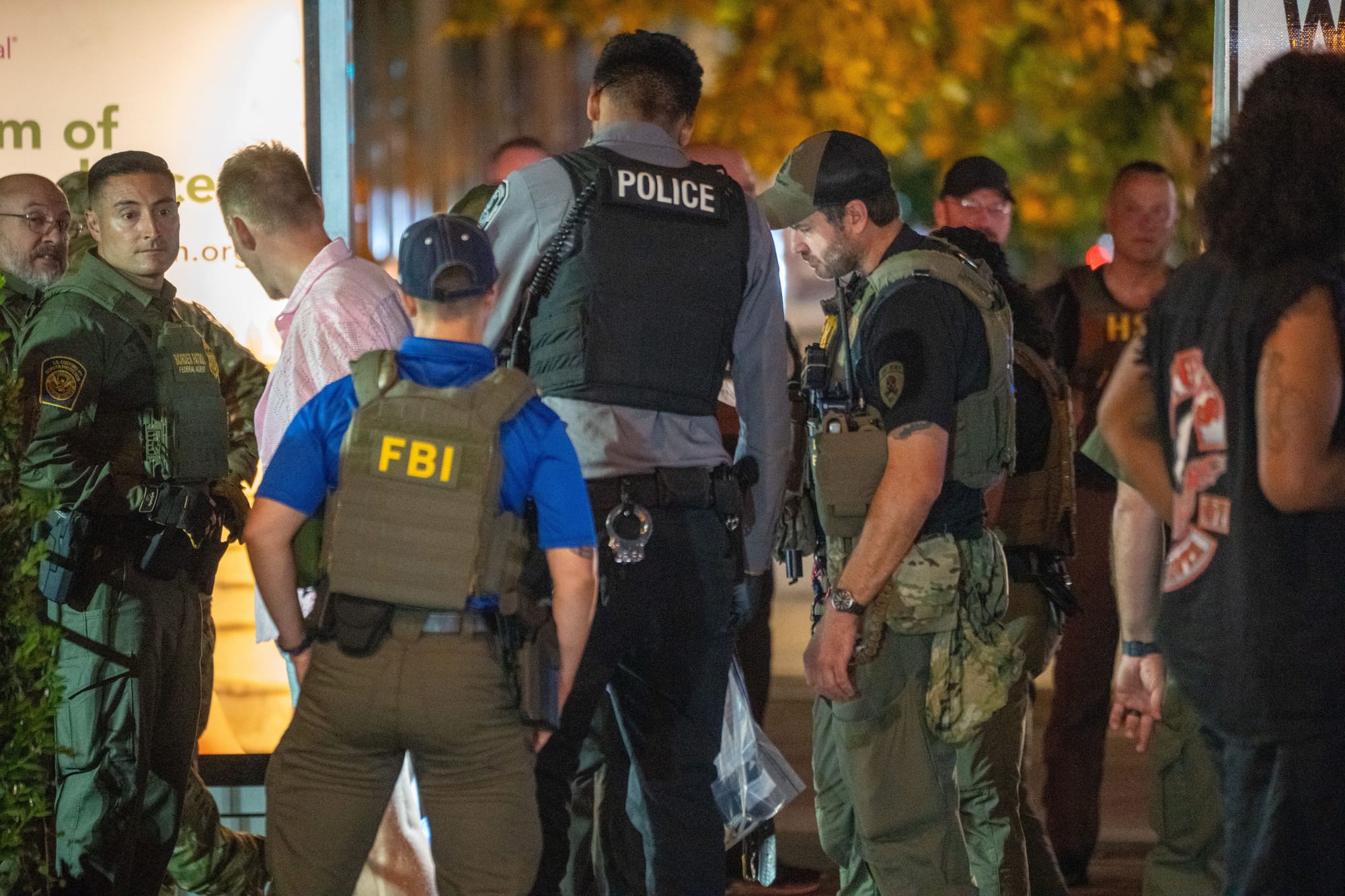 Groups of law enforcement officers—some wearing “FBI” tactical vests, others labeled “POLICE” or in military-style gear—stand together on a nighttime street, engaged in coordination or briefing.