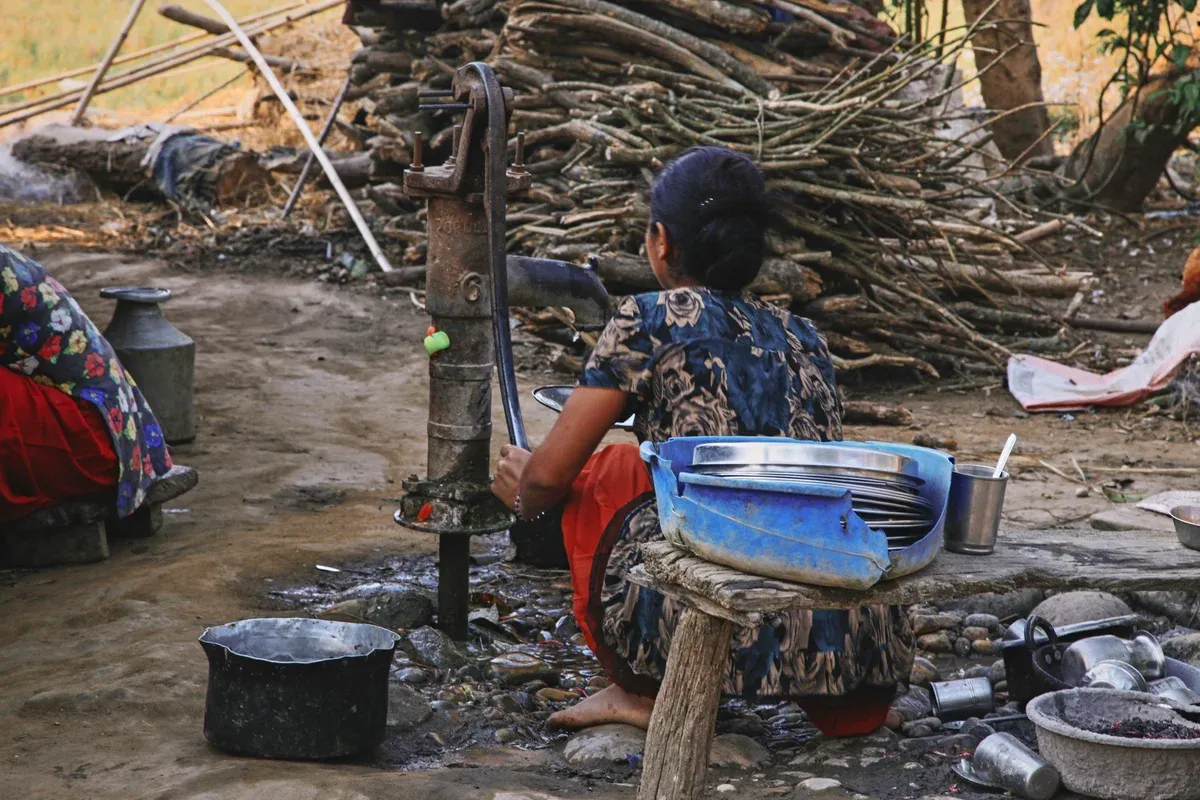 A rural woman operates an iron hand pump to draw water, using it to wash a stack of metal dishes beside her in an outdoor, rustic setting.