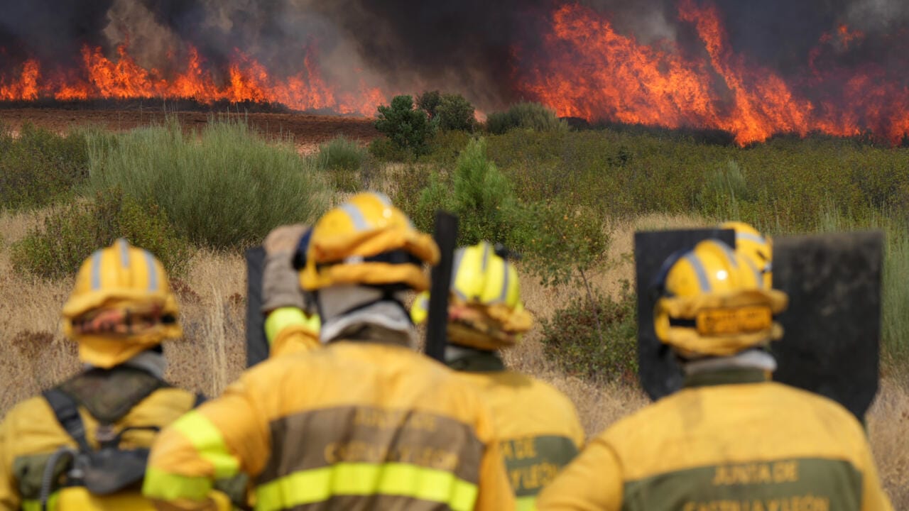 Firefighters in yellow protective gear stand in the foreground while a large wildfire burns through dry vegetation in the background, with flames and thick smoke rising.