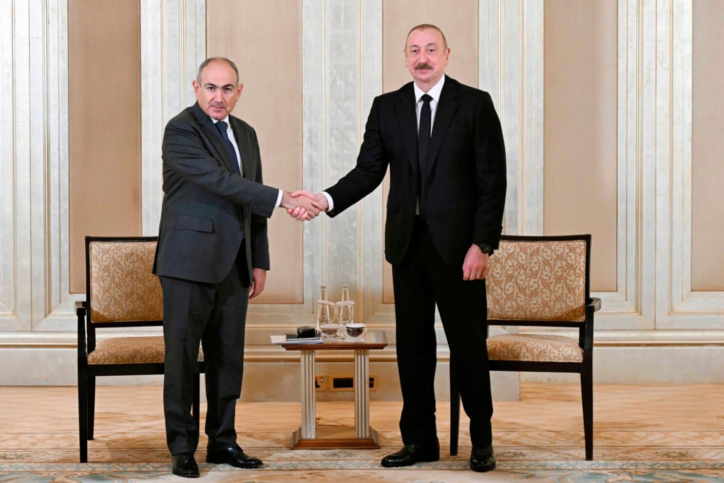 Armenian Prime Minister Nikol Pashinyan and Azerbaijani President Ilham Aliyev shake hands during a diplomatic meeting in a formal setting, with two chairs and a small table between them.