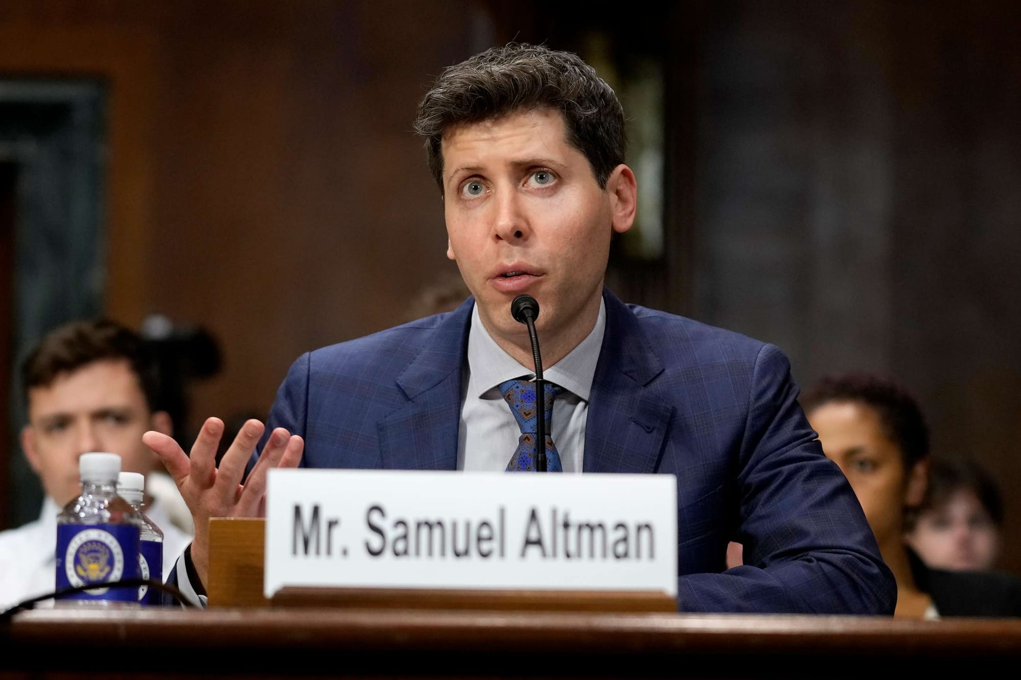 Sam Altman, OpenAI CEO, seated at a hearing desk behind a label reading “Mr. Samuel Altman,” gesturing with one hand while speaking into a microphone during a congressional hearing.