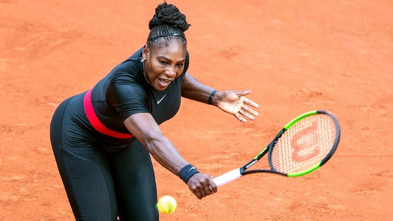 Serena Williams in action on a clay court, stretching forward to return a shot while wearing a black Nike catsuit with a red waistband.