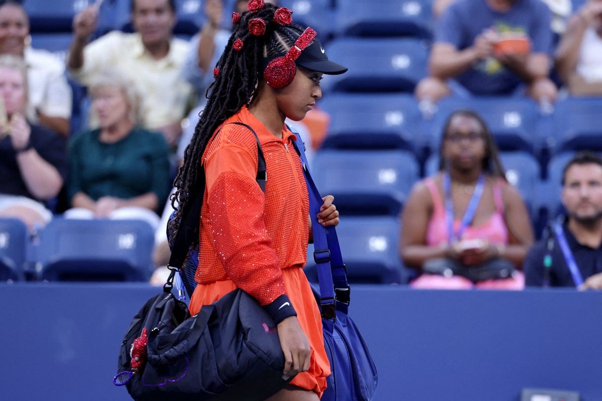 Female tennis player in a bright red outfit with braided hair and red headphones walks onto the court carrying two sports bags, while spectators watch in the background.
