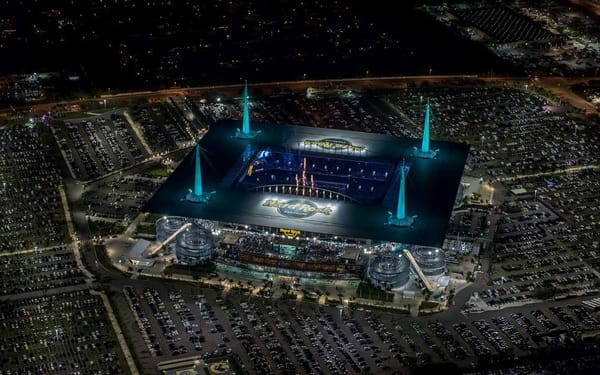 Aerial night view of Hard Rock Stadium filled with fans and cars, lit in vibrant blue.