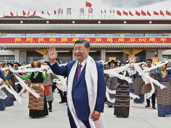 A political leader waves while surrounded by people in traditional Tibetan clothing during a celebration in Lhasa, Tibet, with Chinese flags and banners in the background.