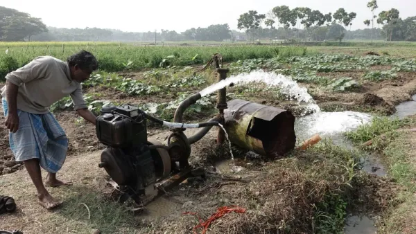 A man leans over a motorized water pump in a rural field, overseeing water flow being directed into nearby furrows or storage trenches.