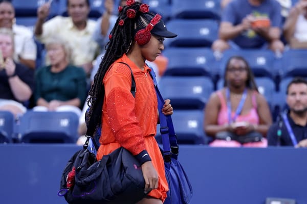 Female tennis player in a bright red outfit with braided hair and red headphones walks onto the court carrying two sports bags, while spectators watch in the background.